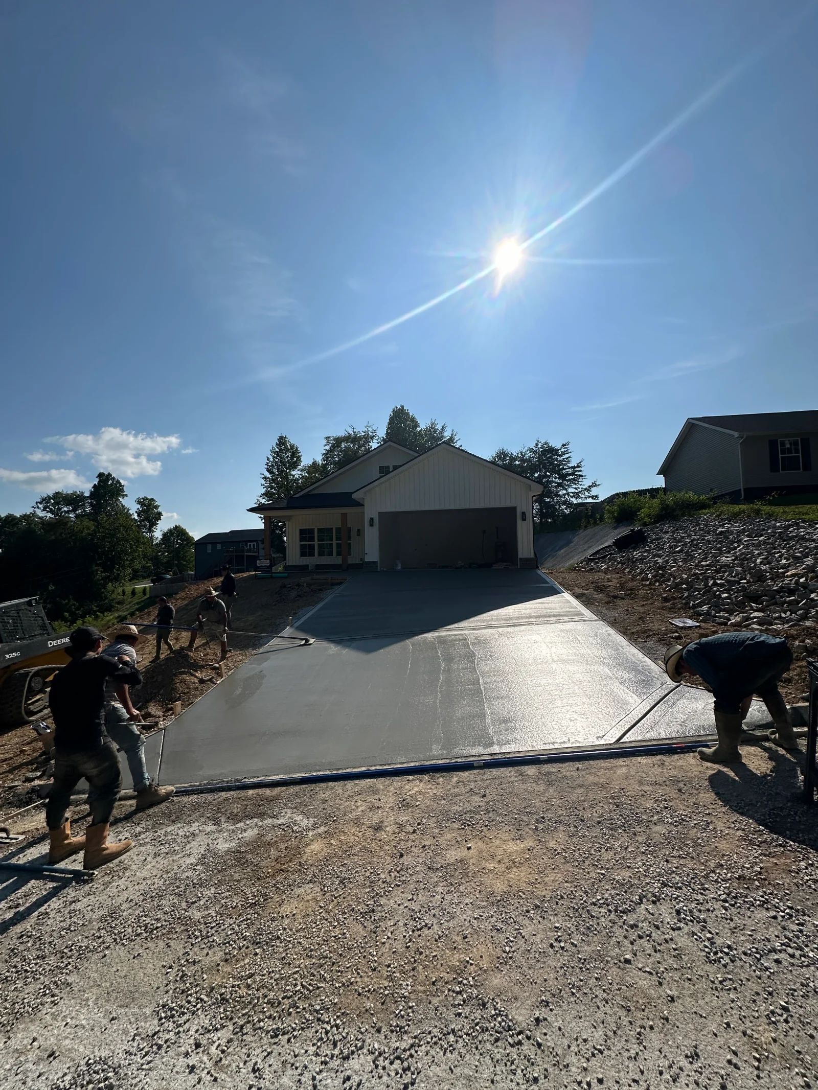 Crew finishing a fresh concrete driveway pour with screeds and bull floats over gravel base beside a new two-car garage