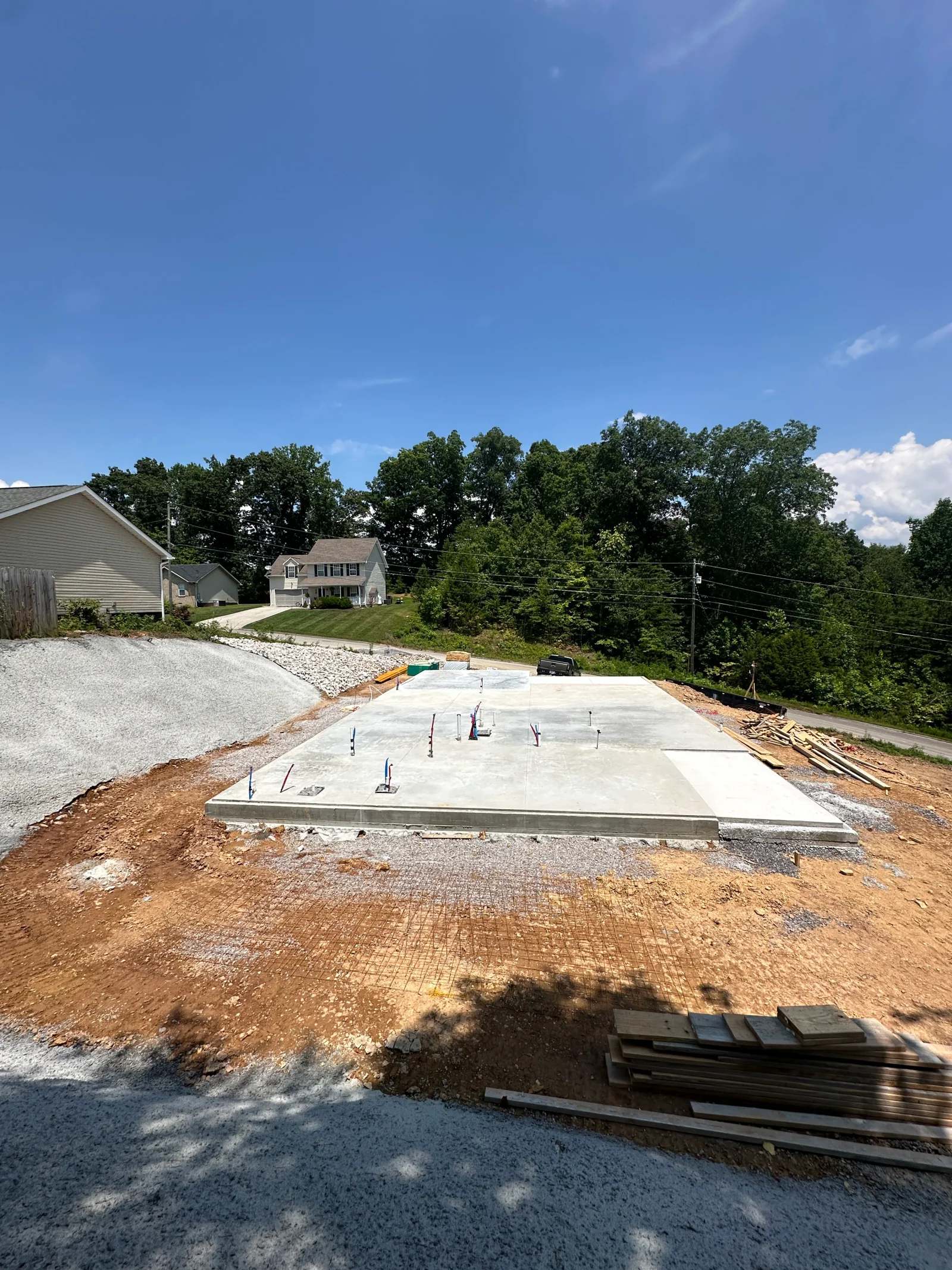 Newly poured concrete slab with rebar anchor bolts and wire mesh on graded clay soil, stacked lumber nearby