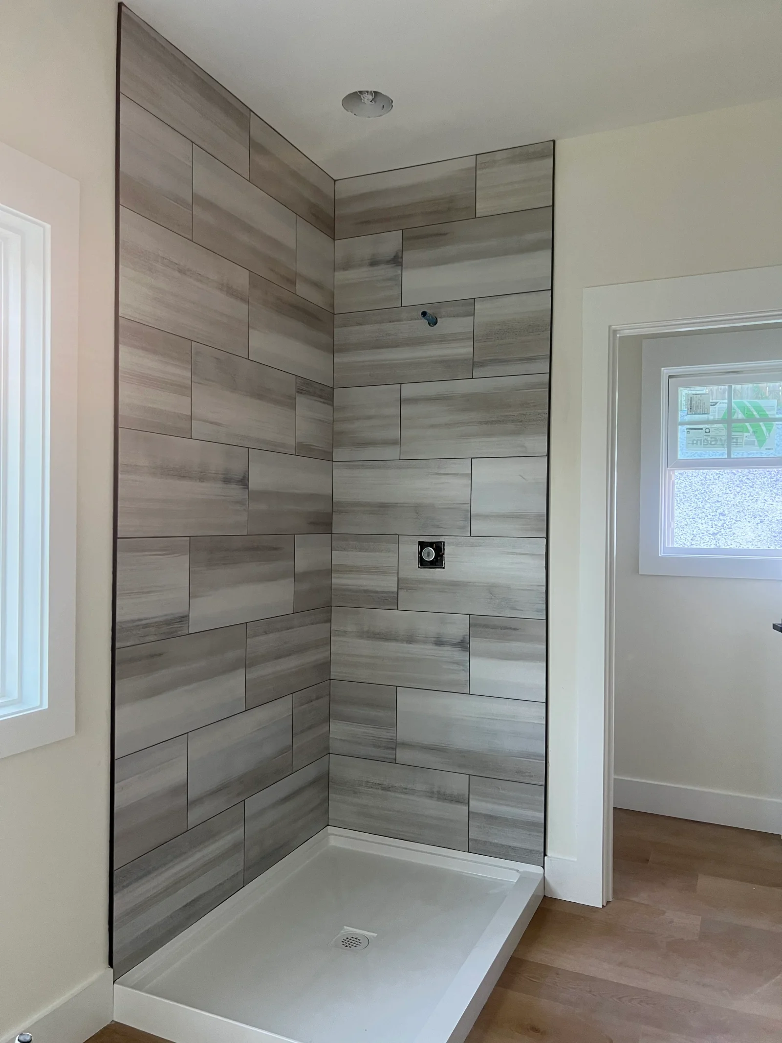 Corner shower with gray wood-look porcelain tile in staggered running bond, white shower pan, and square shower valve trim