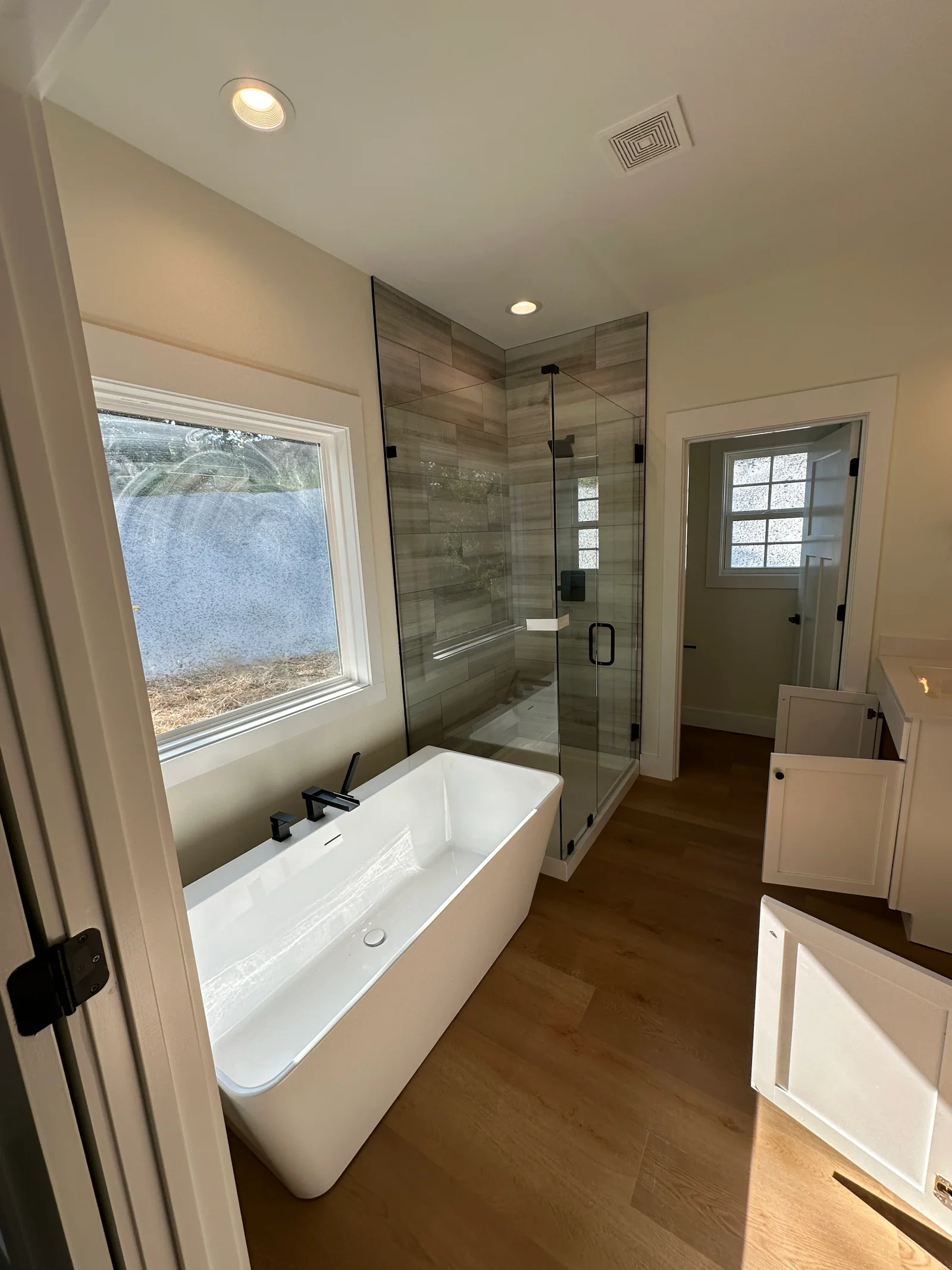 Remodeled bathroom with freestanding white tub, black fixtures, wood-look tile shower with glass enclosure, and white shaker cabinets
