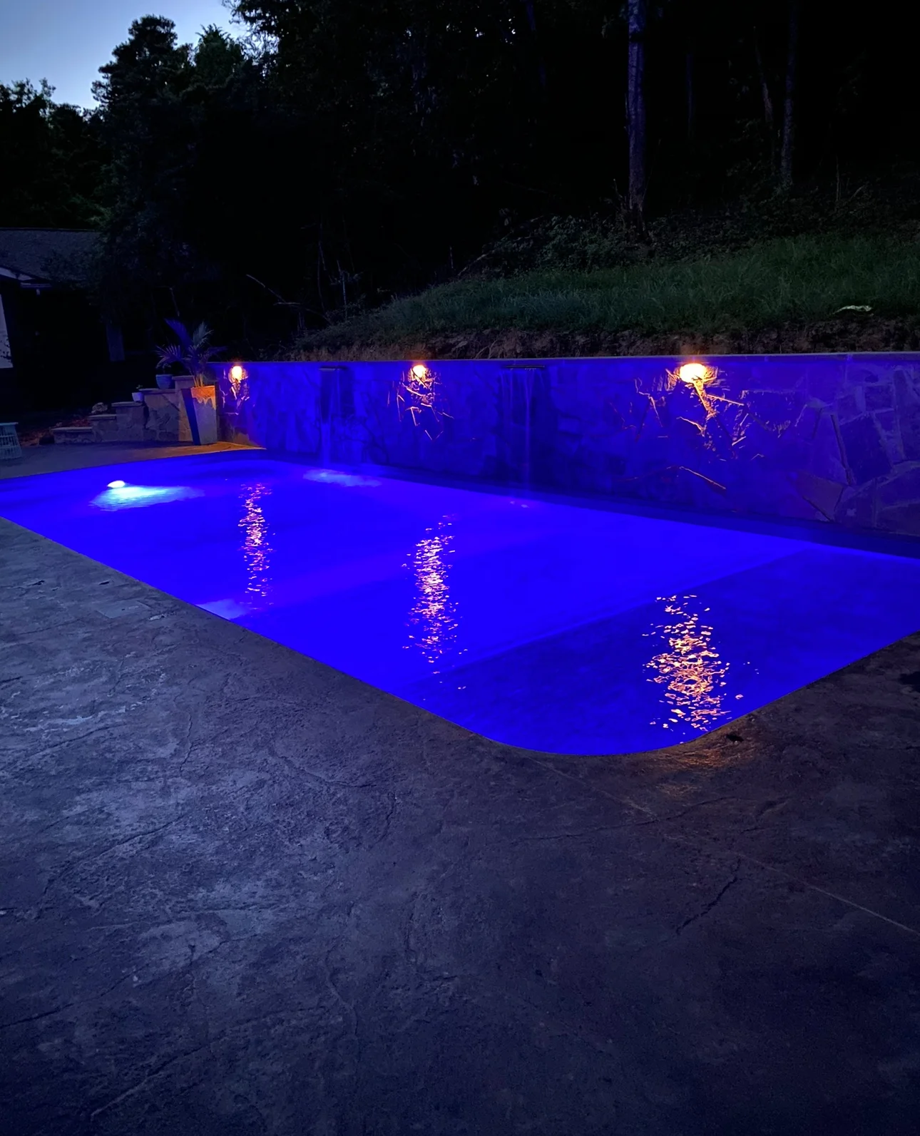 Illuminated in-ground pool at dusk with blue underwater lights, stone veneer wall with wall sconces, and stamped concrete deck
