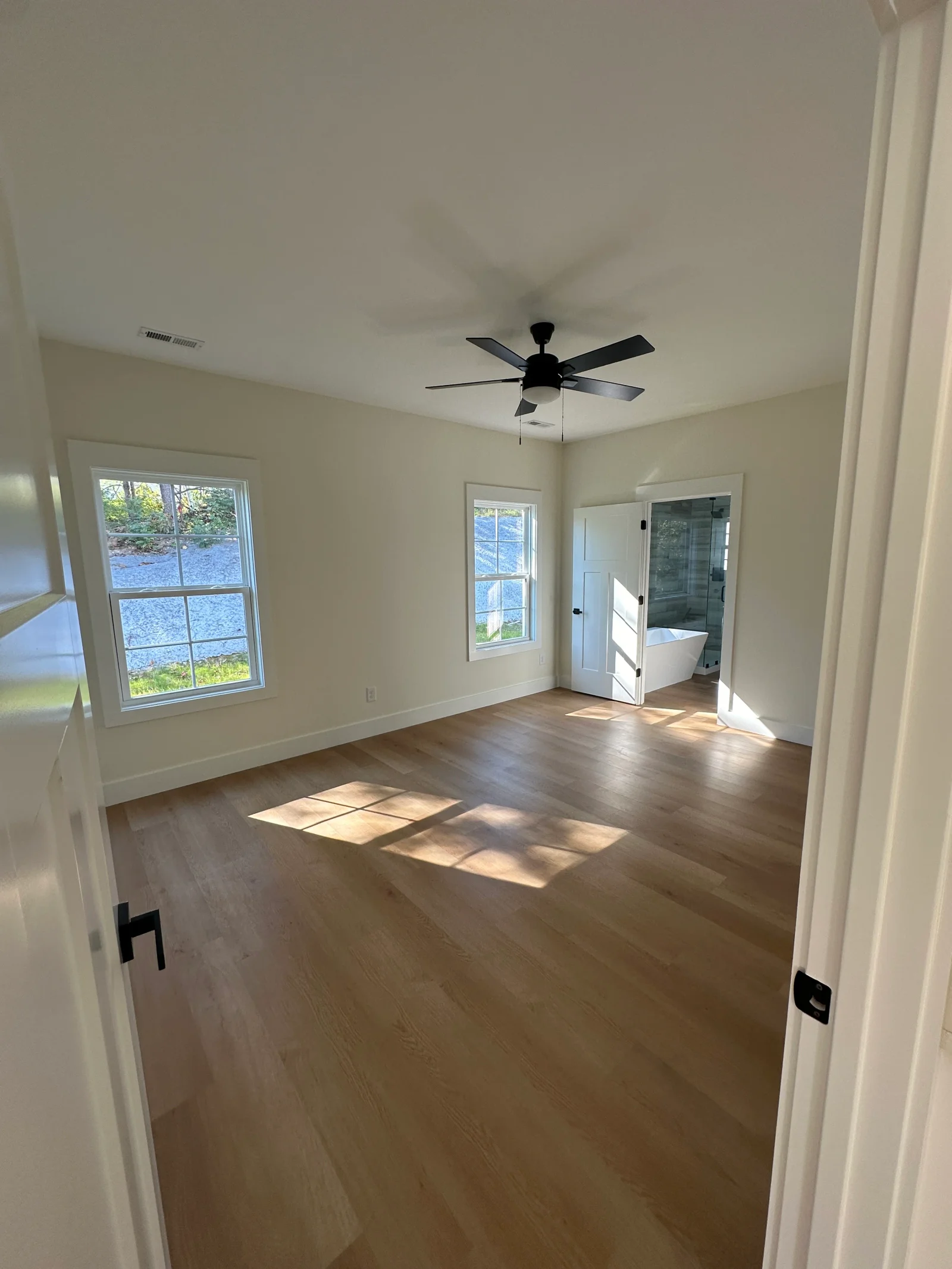 Empty bedroom with wood-look vinyl plank floors, white trim, black ceiling fan, and ensuite with freestanding tub