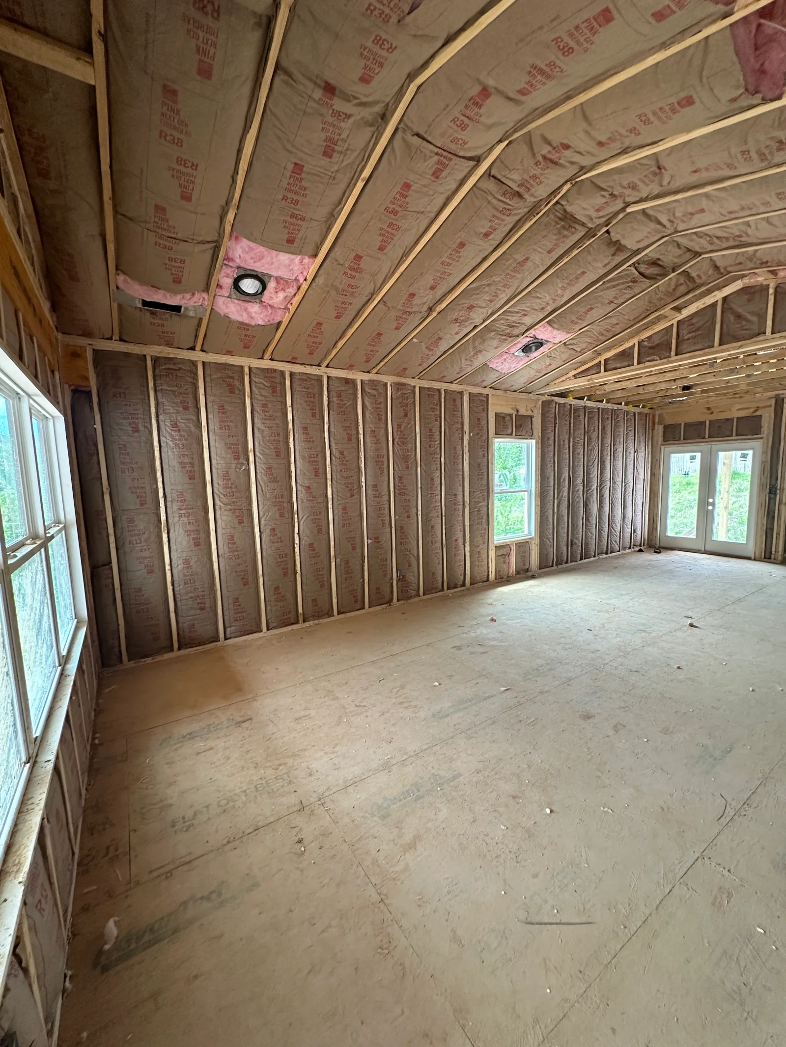 Interior framed room with pink fiberglass insulation in vaulted ceiling and walls, exposed wood studs, and installed windows and French doors.