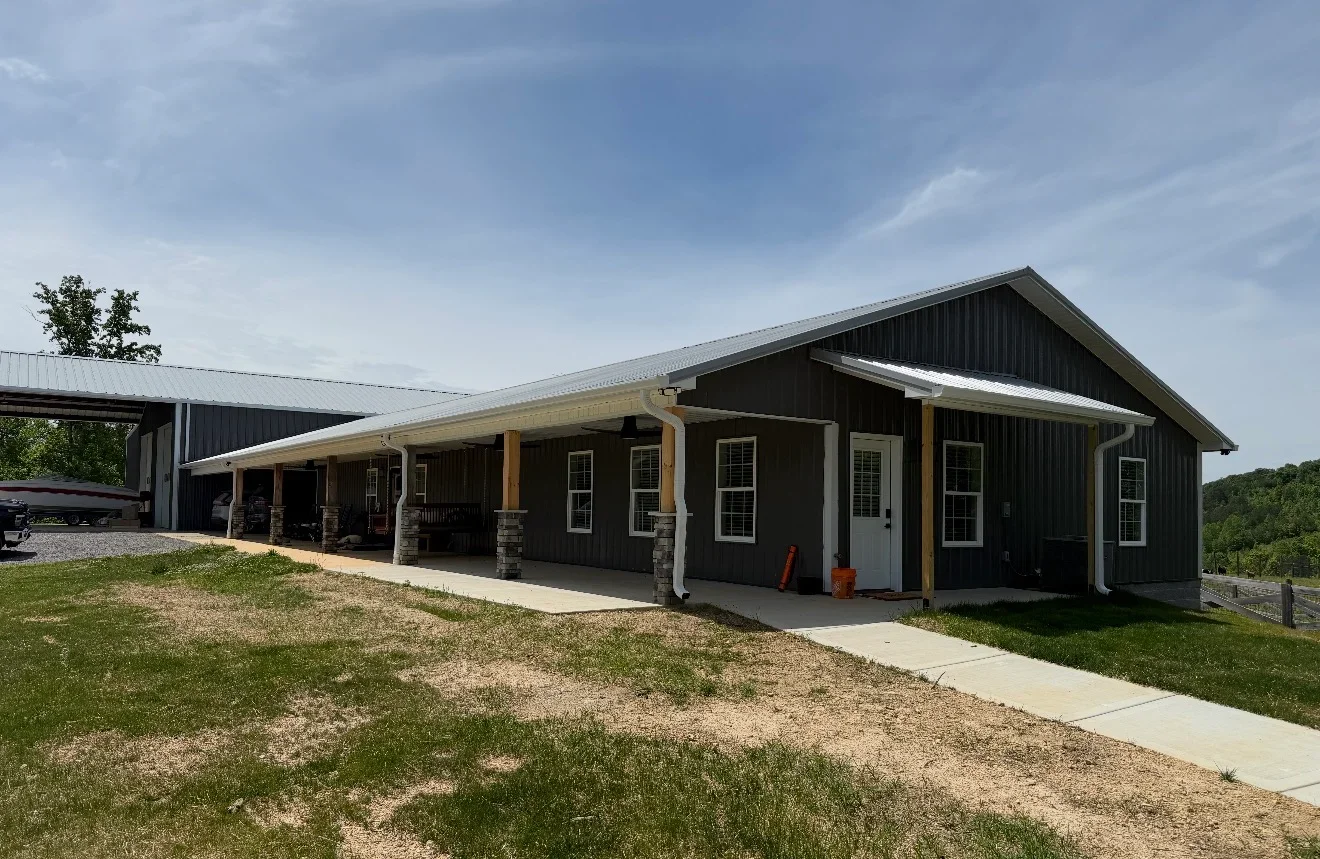 Dark gray metal-sided ranch home with white trim, standing-seam metal roof, wood porch posts on stone bases, and concrete porch.