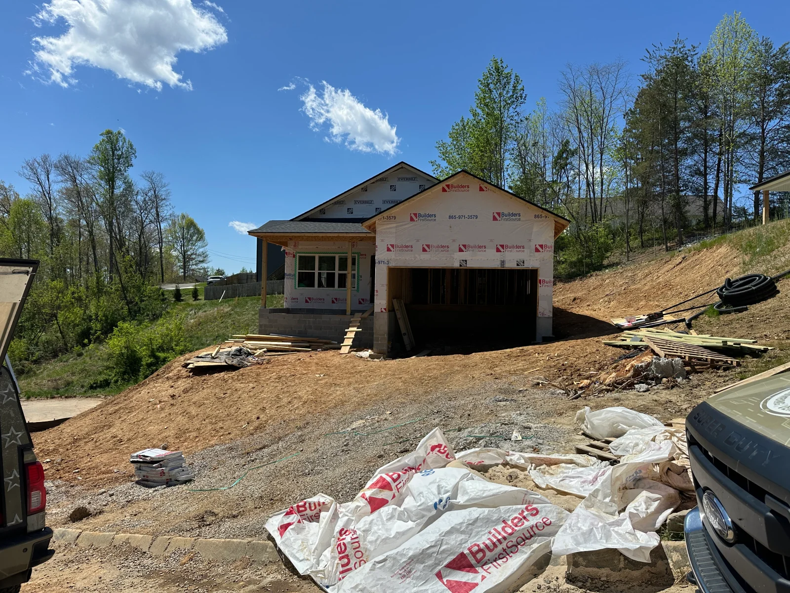 Framed house under construction with house wrap, block foundation, open garage bay, and stacked lumber on sloped site