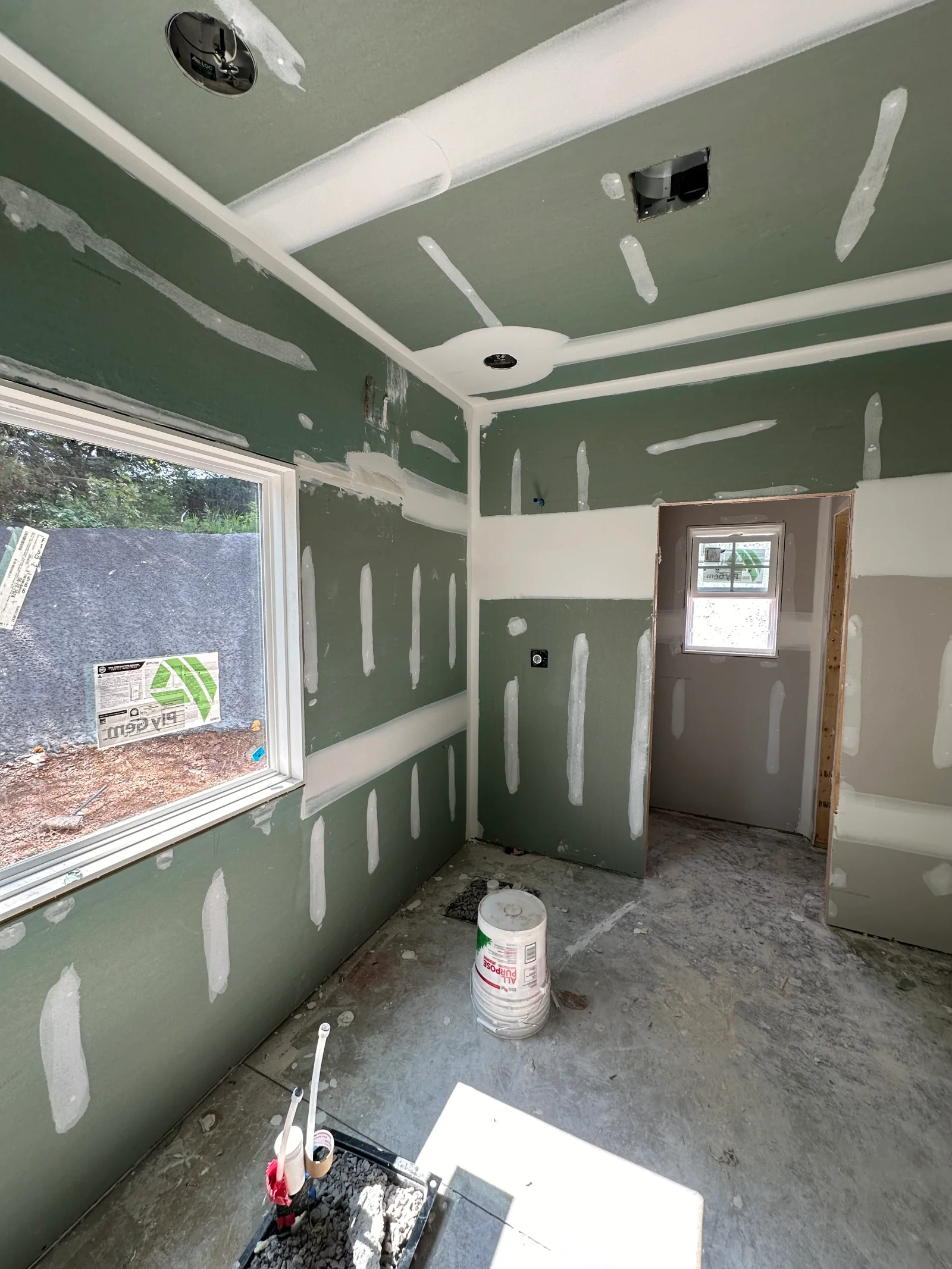 Green moisture-resistant drywall with joint compound on seams, white vinyl window, recessed ceiling lights, and concrete floor during interior finish work