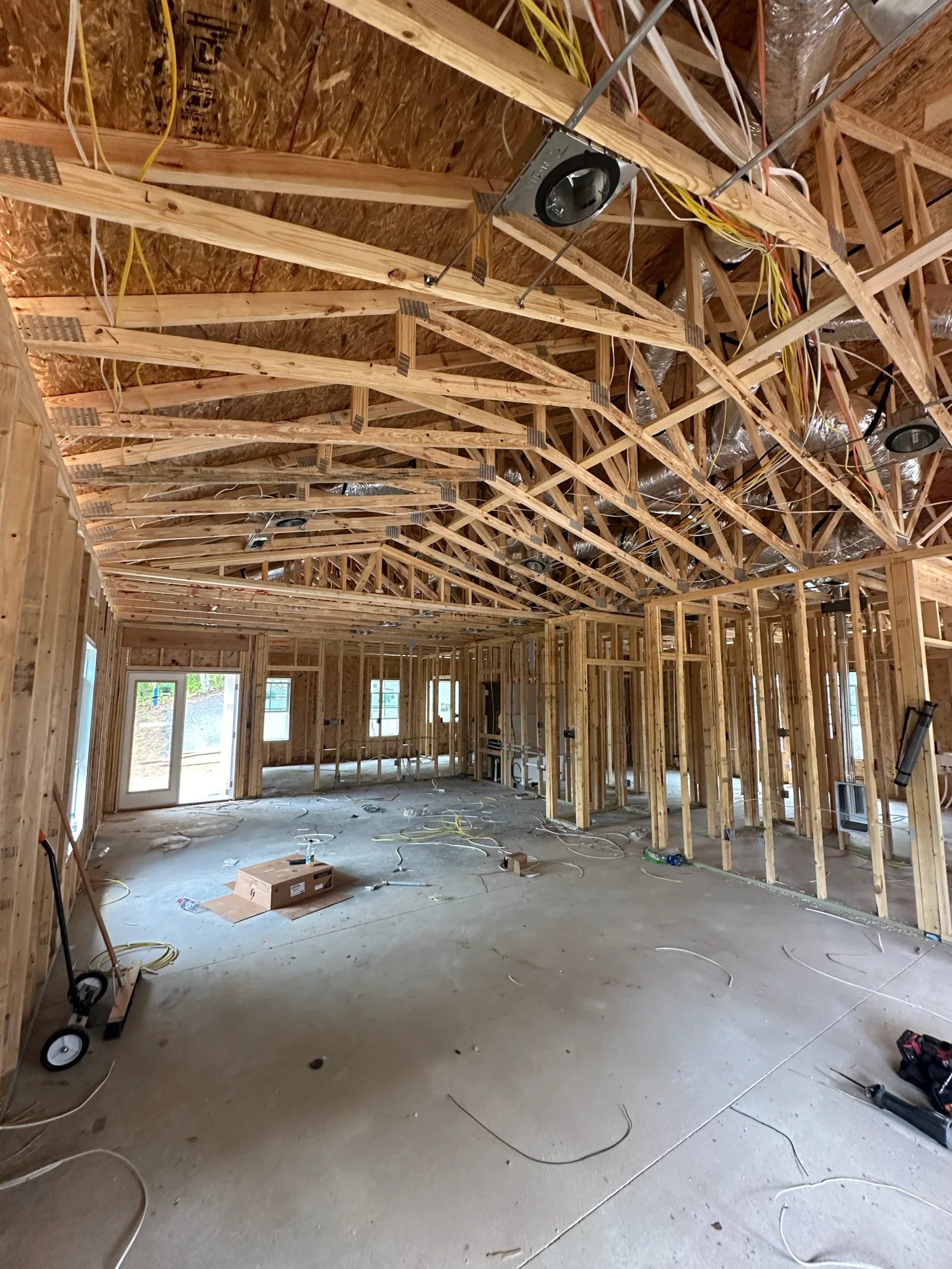 Open interior framing with wood stud walls, pitched wood truss roof, recessed light housings, wiring, and ductwork over a bare concrete slab