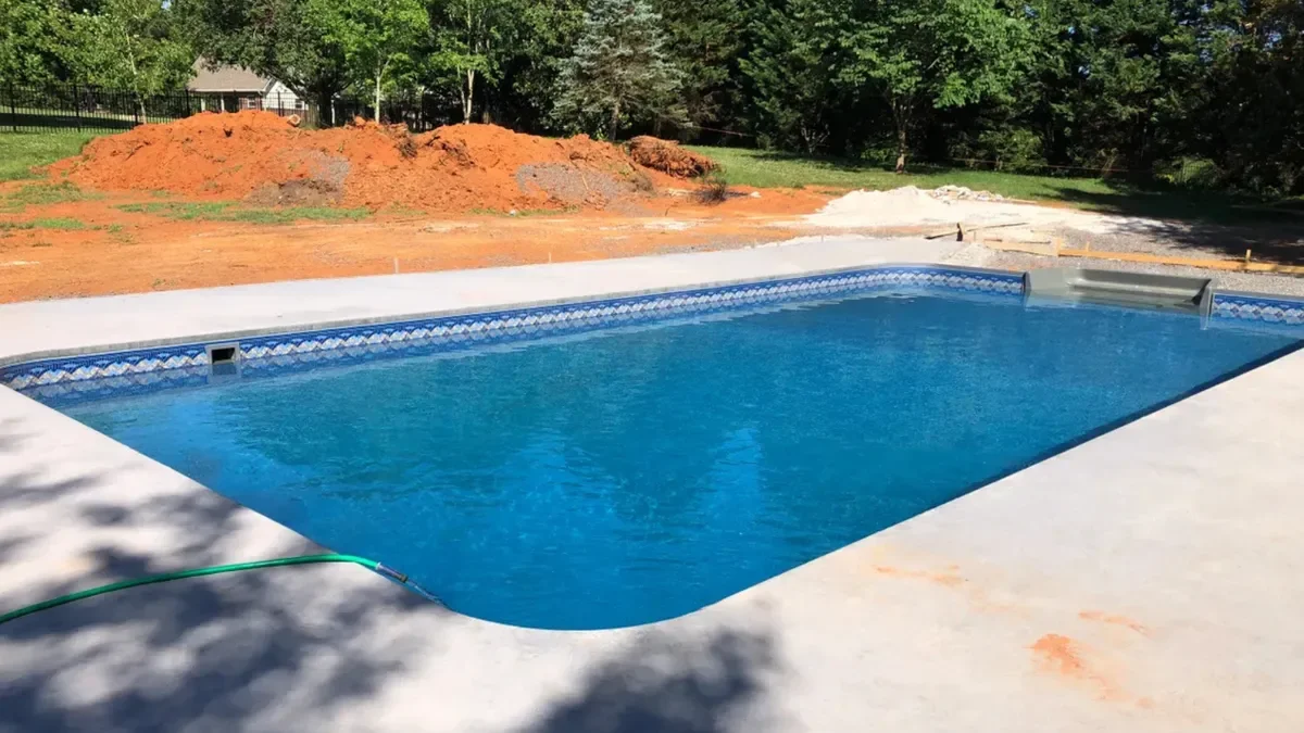 Inground pool with blue and white patterned tile border and light gray concrete deck, under construction