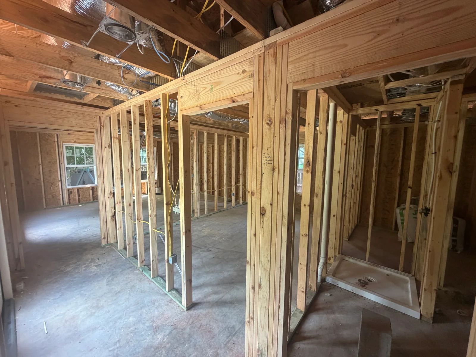 Interior wood stud wall framing with yellow electrical wire, PVC drain pipe, and acrylic shower pan on concrete floor