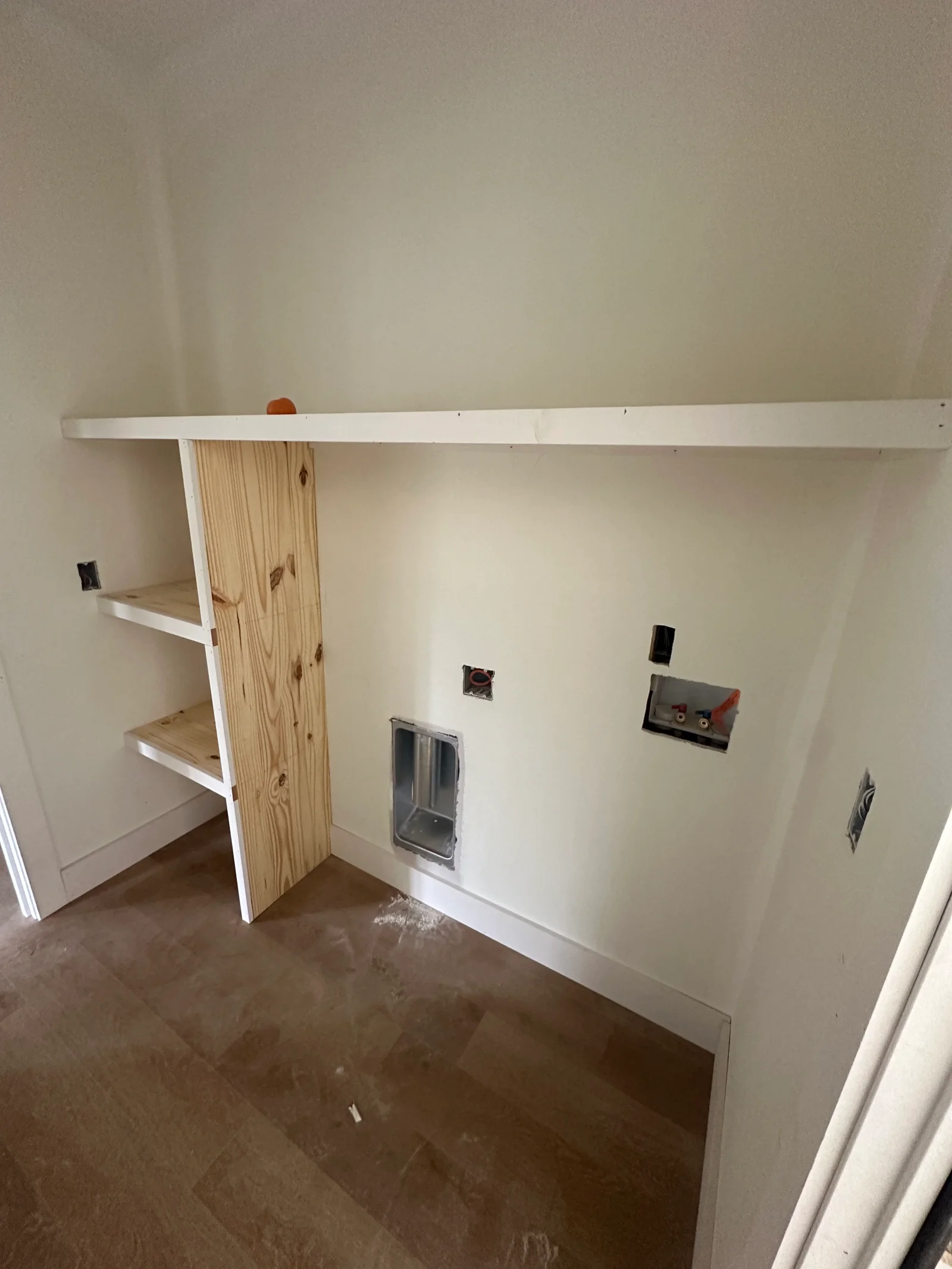 Laundry room under construction with white painted top shelf, pine side shelving, washer water valve box, drain standpipe, and dryer vent rough-in