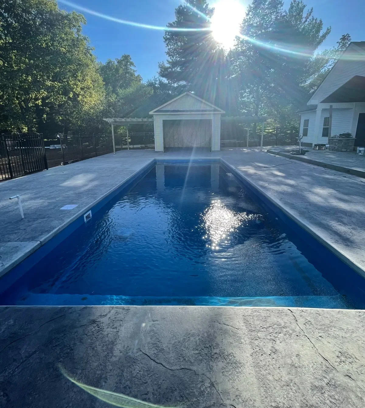 Rectangular inground pool with blue interior and stamped gray concrete deck, black metal fencing, and white pergola and pool house