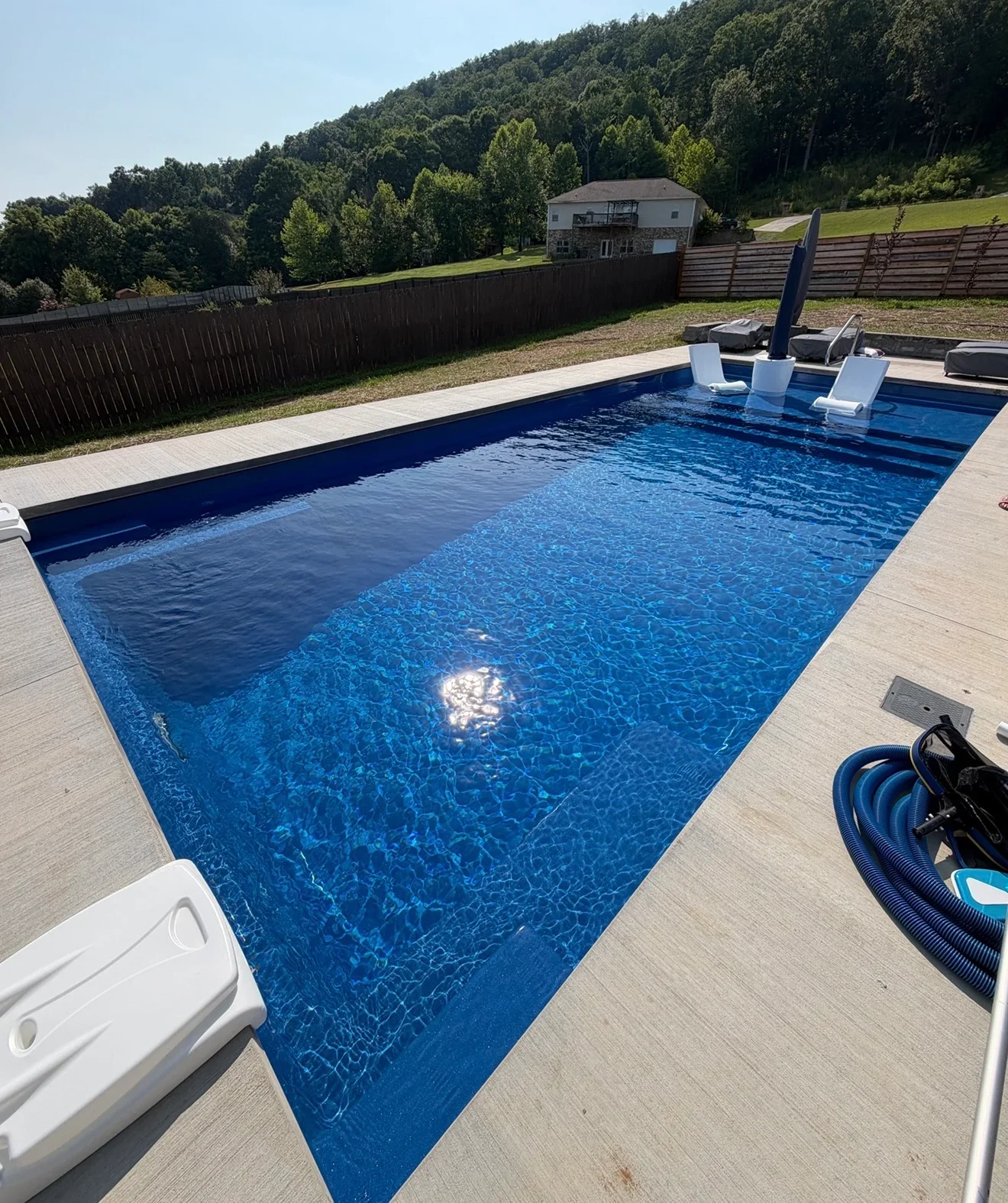 In-ground fiberglass pool with blue shell, light gray concrete deck, white loungers and blue pool hose