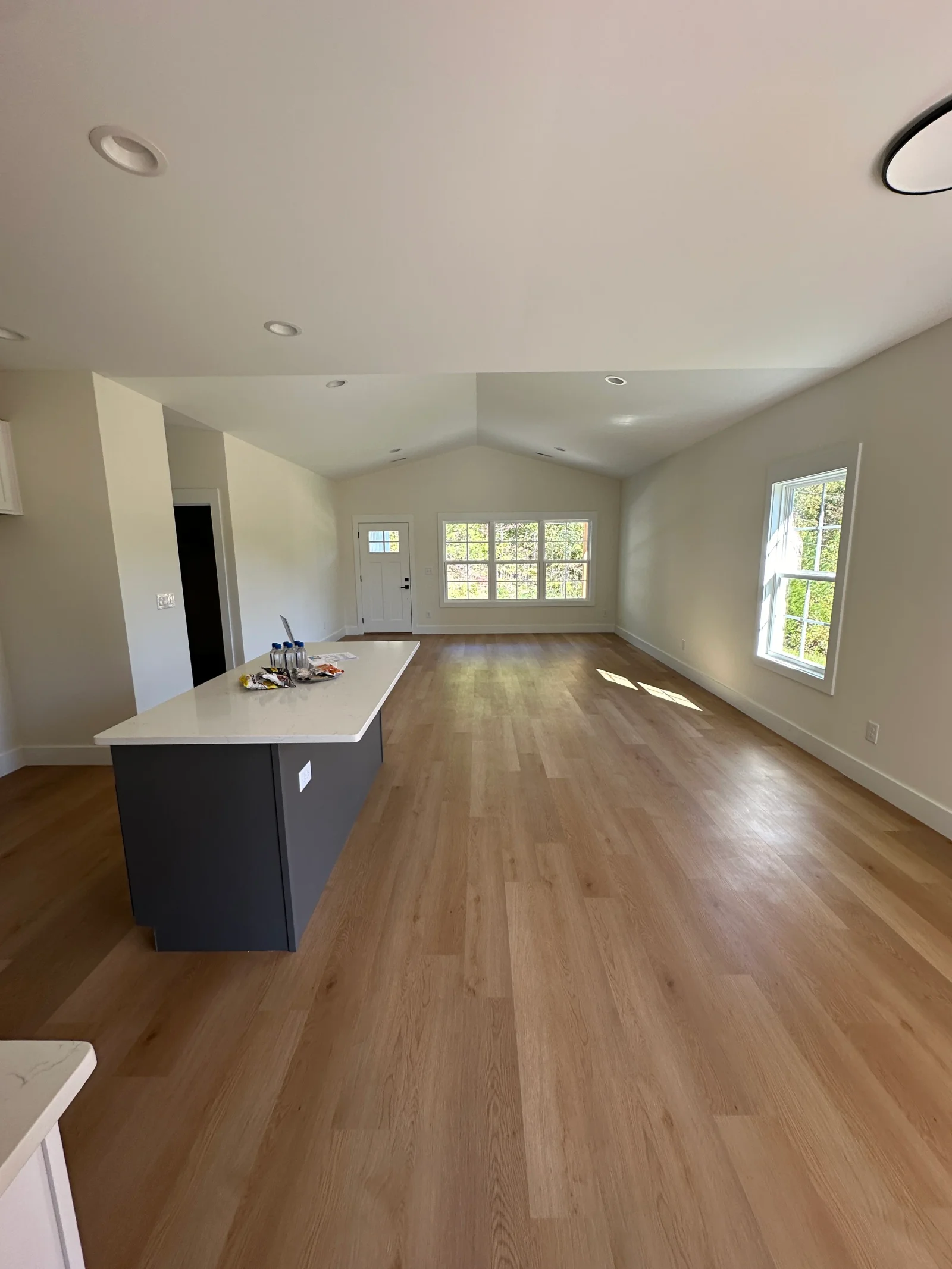 Open living room with vaulted ceiling, wood-look vinyl plank floors, gray island with white quartz countertop, and white-trimmed windows